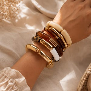 May include: A stack of five bracelets featuring a variety of textures and colors. The set includes polished gold-tone metal tubes, a smooth cream-colored resin piece, a dark brown translucent resin link, and a leopard print patterned segment.