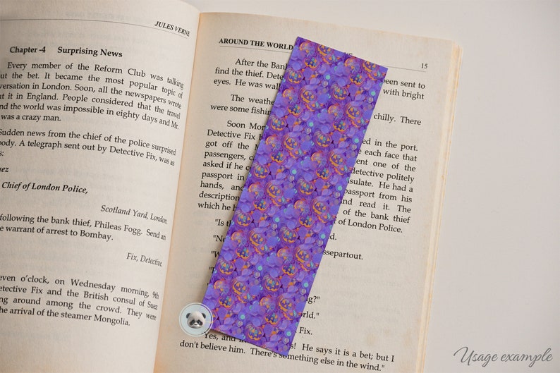 May include: A purple and orange bookmark with a repeating pattern of jack-o'-lantern faces. The bookmark is made of a soft, textured material and has a slightly glittery finish.