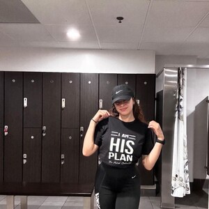 Puede incluir: Una persona con una camiseta negra con el texto "I AM HIS PLAN" en blanco. La persona lleva una gorra de b&eacute;isbol negra y pantalones negros. La persona est&aacute; de pie frente a una fila de taquillas.