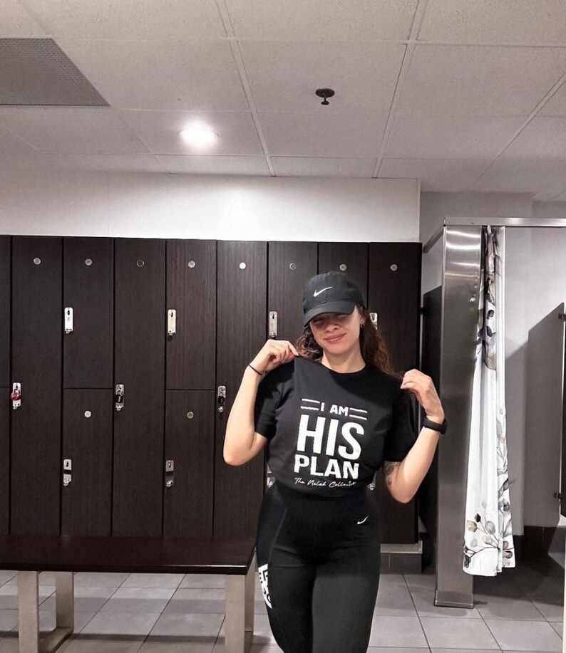 Puede incluir: Una persona con una camiseta negra con el texto "I AM HIS PLAN" en blanco. La persona lleva una gorra de b&eacute;isbol negra y pantalones negros. La persona est&aacute; de pie frente a una fila de taquillas.