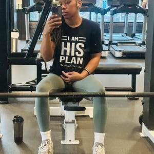 Puede incluir: Una persona con una camiseta negra con el texto "I AM HIS PLAN" en letras blancas. La persona est&aacute; sentada en un banco de pesas en un gimnasio.