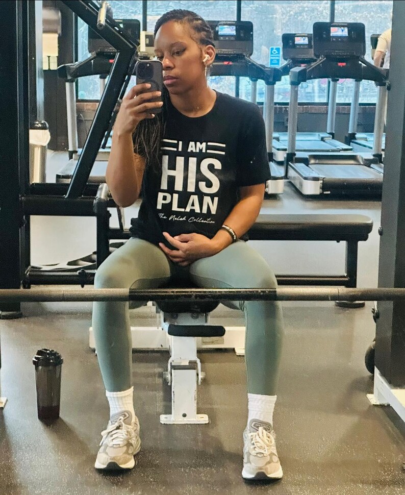 Puede incluir: Una persona con una camiseta negra con el texto "I AM HIS PLAN" en letras blancas. La persona est&aacute; sentada en un banco de pesas en un gimnasio.