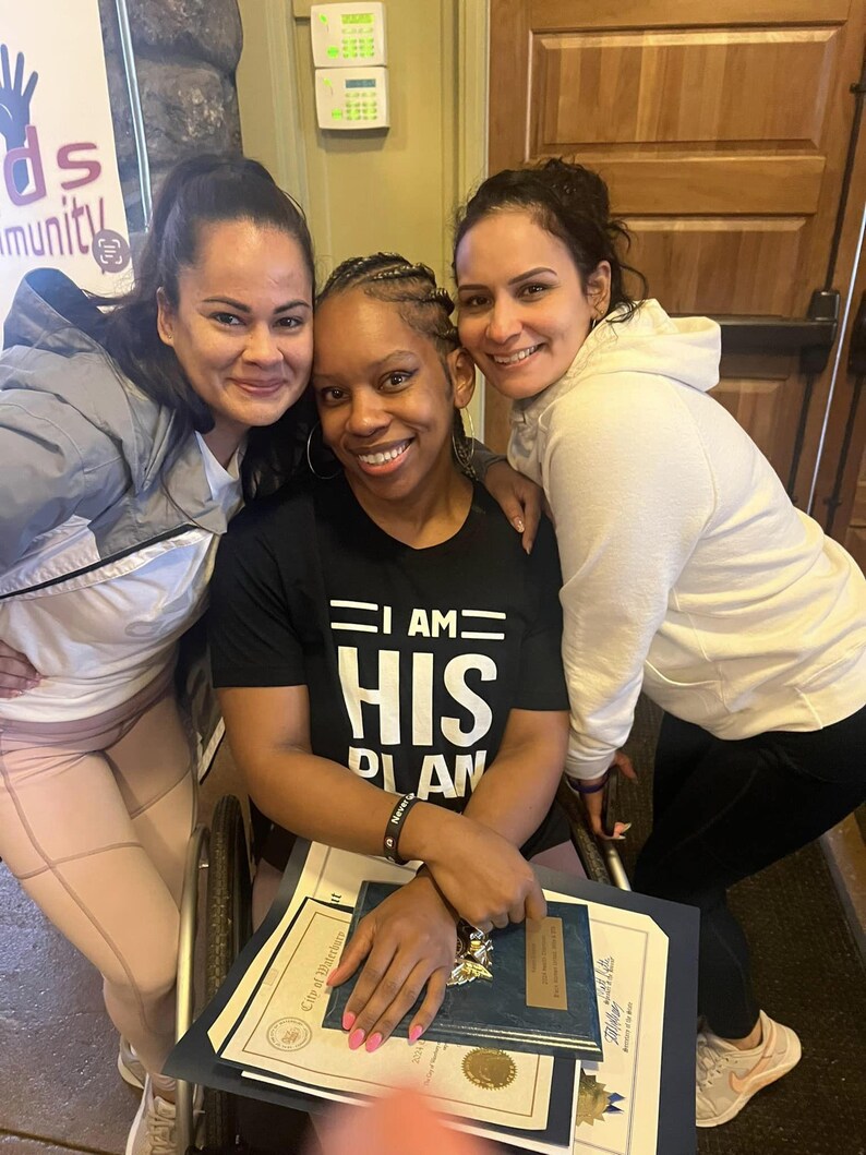 Puede incluir: Tres mujeres sonriendo juntas. La mujer del medio est&aacute; sentada en una silla de ruedas y lleva una camiseta negra que dice "I AM HIS PLAN". Las mujeres sostienen certificados y premios.