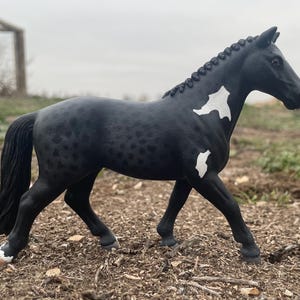 May include: A black horse figurine with white markings on its body. The horse has a braided mane and tail. The figurine is standing on a bed of brown mulch. The horse's hooves are black with white accents.