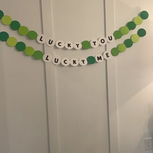 May include: A decorative banner with green and light green circles, spelling out "LUCKY YOU" and "LUCKY ME" in black letters on white circles. The banner is hanging against a white wall.
