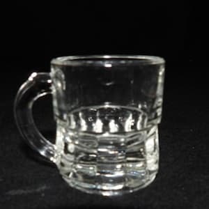 May include: A small, clear glass mug with a handle. The mug features a textured, patterned design around the base and a smooth, cylindrical upper section. The handle is curved and attached to the side.