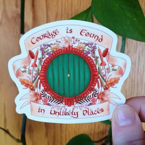 May include: A die-cut sticker featuring a round green door with a central gold knob, framed by a red circular border. The design includes floral and botanical illustrations with a banner displaying the text Courage is Found in Unlikely Places.