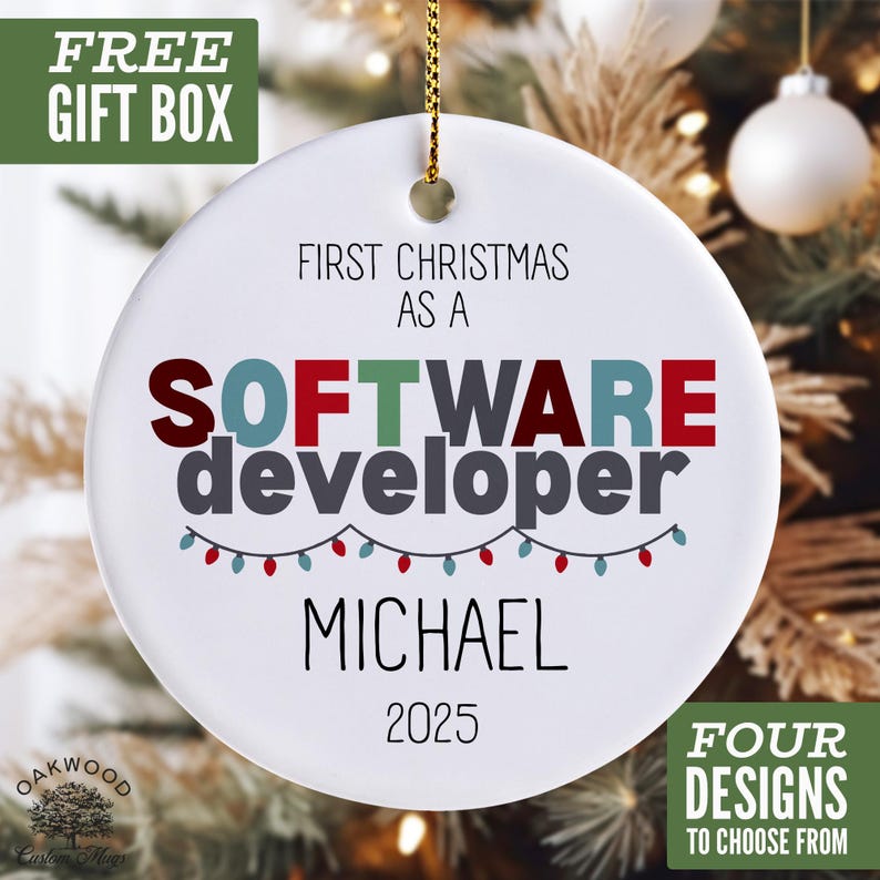 a white ornament with the text "FIRST CHRISTMAS AS A SOFTWARE DEVELOPER MICHAEL 2025" on it, along with a "FREE GIFT BOX" and "FOUR DESIGNS TO CHOOSE FROM" text. The ornament is hanging in front of a Christmas tree, creating a festive and personalized holiday decoration.