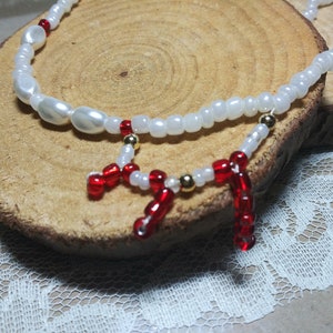 May include: A white and red beaded necklace with a large white pearl and gold accents. The necklace is displayed on a wooden surface.
