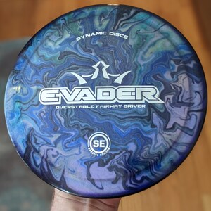 May include: A blue, black and green disc golf disc with a swirl pattern. The disc is labelled "Evader" and "Overstable Fairway Driver" with a logo that says "Dynamic Discs". The disc also has a "Special Edition" logo.