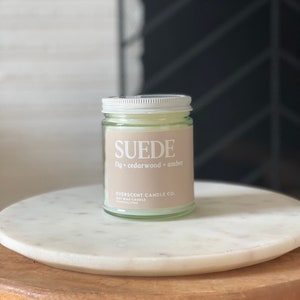 May include: A clear glass candle jar with a white lid. The candle is labeled "SUEDE fig + cedarwood + amber" and "EVERSCENT CANDLE CO. SOY WAX CANDLE HAND-POURED".