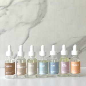 May include: Six glass bottles with white droppers, each labeled with a different scent: Santal, Coco Cashmere, Suede, Ocean, Surf, Baja Flora, and Summer. The bottles are filled with a clear liquid and sit on a white surface against a marble background.