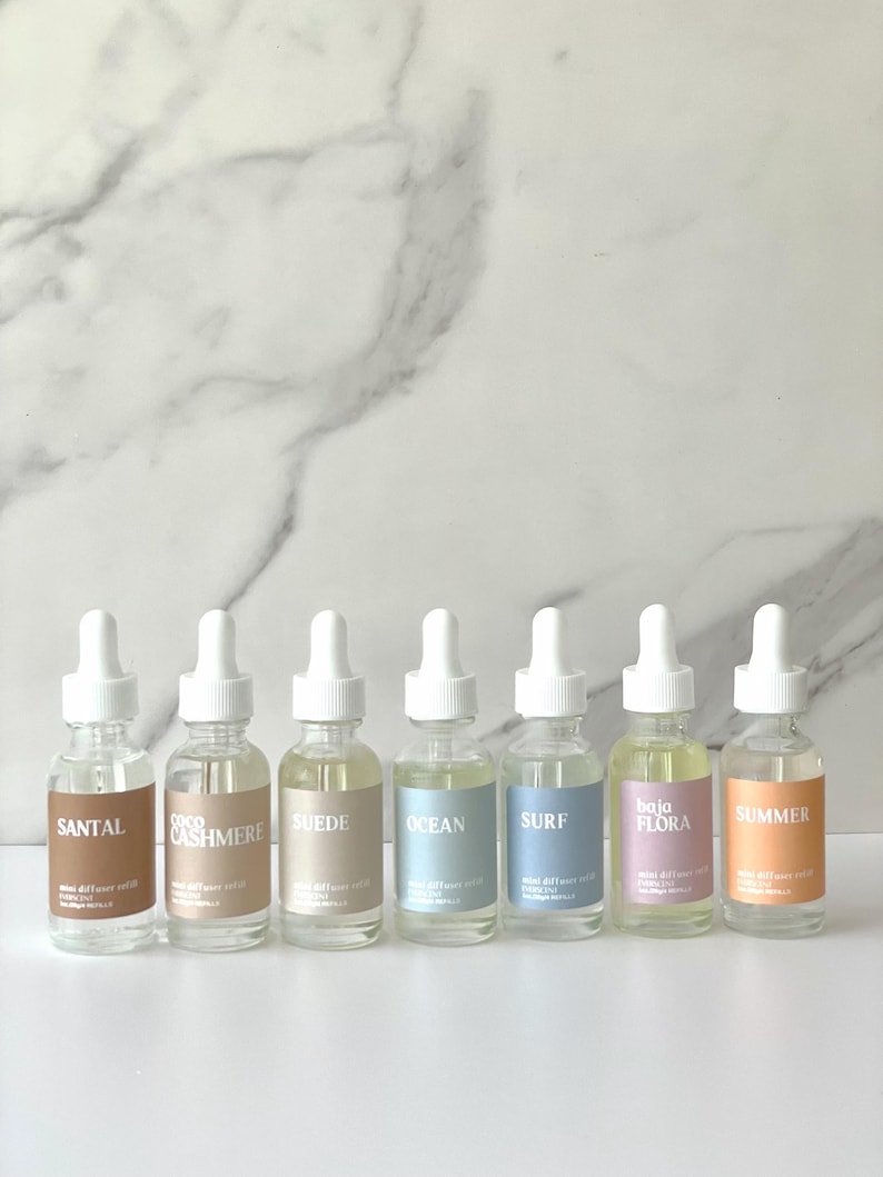 May include: Six glass bottles with white droppers, each labeled with a different scent: Santal, Coco Cashmere, Suede, Ocean, Surf, Baja Flora, and Summer. The bottles are filled with a clear liquid and sit on a white surface against a marble background.