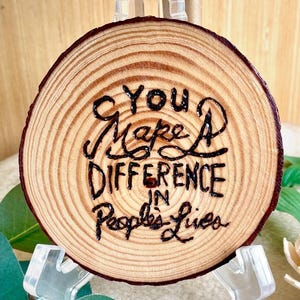 May include: A circular wooden slice featuring visible tree rings and a dark, hand-burned inscription that reads: You Make A DIFFERENCE IN People's Lives. The natural light brown wood grain is prominent, and the edges are dark brown and textured.
