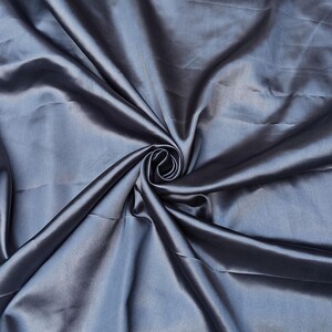 Puede incluir: Primer plano de una tela plegada y brillante en un tono azul plateado apagado. El material est&aacute; drapeado y arremolinado, creando un patr&oacute;n de sombras y luces. El brillo de la tela sugiere una textura satinada o similar a la seda.
