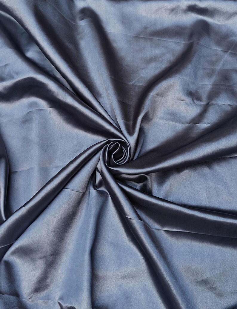 Puede incluir: Primer plano de una tela plegada y brillante en un tono azul plateado apagado. El material est&aacute; drapeado y arremolinado, creando un patr&oacute;n de sombras y luces. El brillo de la tela sugiere una textura satinada o similar a la seda.