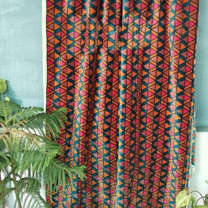 以下が含まれることがあります： 幾何学模様のカーテン。オレンジ、ピンク、ティール色の三角形の繰り返し模様が特徴です。カーテンは水色の壁の前に吊り下げられており、手前に植物があります。