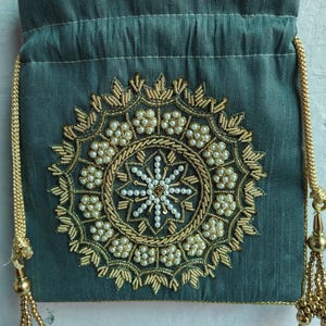 Könnte beinhalten: Eine tealgrüne Kordeltasche mit einem goldenen und weißen Perlenmuster in Kreisform. Die Tasche hat goldene Quasten.