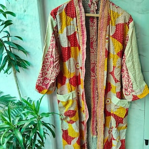 Könnte beinhalten: Ein farbenfroher Kimono-Stil-Bademantel mit einem Patchwork-Design in Rot, Gelb und Creme. Der Bademantel hat Taschen und hängt an einem Holzbügel. Die Ärmel sind dreiviertellang und der Bademantel ist aus leichtem Stoff.