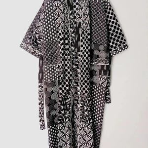May include: A black and white patterned robe with a patchwork design. The robe features various geometric and floral patterns, including polka dots, swirls, and leaf motifs. It has a tie belt and is hanging on a hanger.