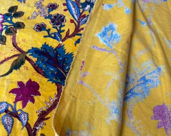 Puede incluir: Tela amarillo brillante con un estampado floral en tonos morados, azules y marrones. El diseño presenta grandes flores y follaje detallado. La tela está doblada, revelando un reverso amarillo más claro.