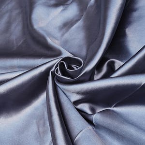 Puede incluir: Primer plano de una tela de sat&eacute;n azul plateado drapeada. El material est&aacute; dispuesto en pliegues y remolinos suaves, creando reflejos y sombras. El brillo de la tela sugiere un textil liso y de alta calidad, ideal para ropa o decoraci&oacute;n del hogar.