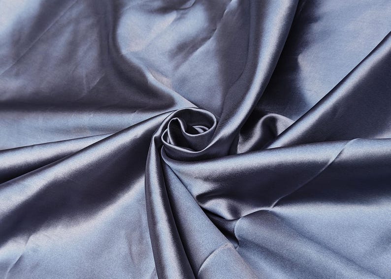 Puede incluir: Primer plano de una tela de sat&eacute;n azul plateado drapeada. El material est&aacute; dispuesto en pliegues y remolinos suaves, creando reflejos y sombras. El brillo de la tela sugiere un textil liso y de alta calidad, ideal para ropa o decoraci&oacute;n del hogar.