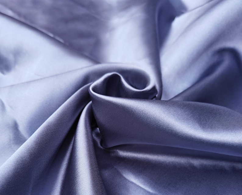 Puede incluir: Primer plano de una tela fluida y lustrosa en un tono suave y apagado de color b&iacute;garo. El material est&aacute; tejido apretado, con l&iacute;neas diagonales visibles, y tiene una textura suave y sedosa. La tela est&aacute; drapeada y doblada, creando sombras y reflejos.