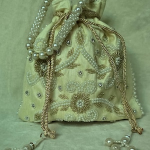 以下が含まれることがあります： アイボリーサテン巾着袋、金と白のビーズで複雑な花の刺繍が施されています。バッグはパールビーズのハンドルとタッセルが特徴で、エレガントで華やかなアクセサリーです。小さなアイテムを持ち運ぶのに最適です。