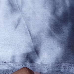 Puede incluir: Primer plano de una tela azul claro con un brillo sedoso. La tela tiene un aspecto texturizado con pliegues y sombras visibles. Un borde cosido con flecos es visible en la parte inferior de la imagen.