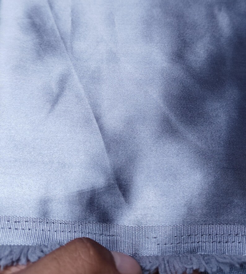 Puede incluir: Primer plano de una tela azul claro con un brillo sedoso. La tela tiene un aspecto texturizado con pliegues y sombras visibles. Un borde cosido con flecos es visible en la parte inferior de la imagen.