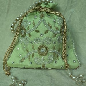 Puede incluir: Un pequeño bolso con cordón de color verde claro con intrincados abalorios y detalles de perlas. El bolso presenta un diseño floral con cuentas doradas y blancas, y un asa de perlas. El cordón es dorado y el bolso tiene borlas de perlas.