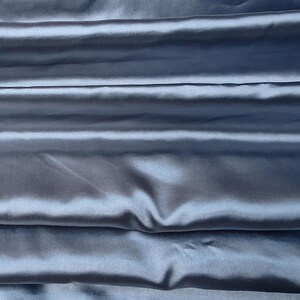Puede incluir: Primer plano de una tela plegada y lustrosa en un tono azul gris&aacute;ceo apagado. El material tiene un brillo sedoso, con pliegues horizontales que crean sombras y reflejos. La tela parece suave y fluida, adecuada para ropa o decoraci&oacute;n del hogar.