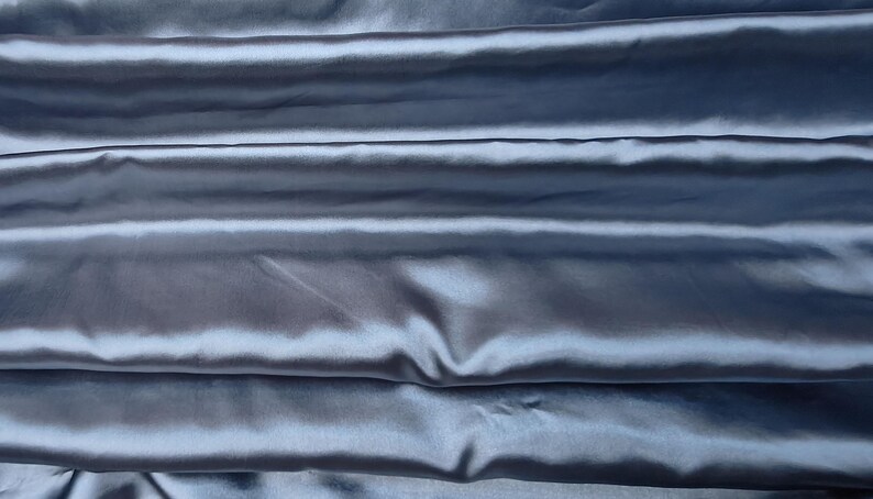 Puede incluir: Primer plano de una tela plegada y lustrosa en un tono azul gris&aacute;ceo apagado. El material tiene un brillo sedoso, con pliegues horizontales que crean sombras y reflejos. La tela parece suave y fluida, adecuada para ropa o decoraci&oacute;n del hogar.
