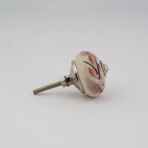 Op de afbeelding: Een witte keramische knop met een bloemenmotief in roze en zwarte tinten. De knop heeft een zilveren schroef en is bevestigd aan een zilveren basis.