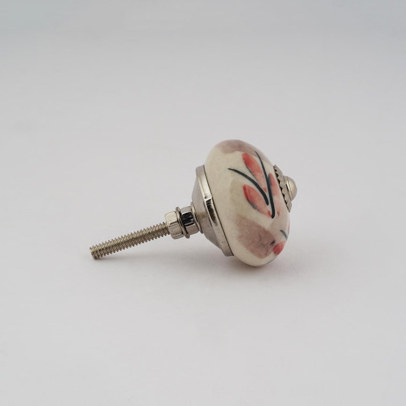 Op de afbeelding: Een witte keramische knop met een bloemenmotief in roze en zwarte tinten. De knop heeft een zilveren schroef en is bevestigd aan een zilveren basis.