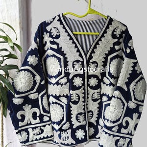 May include: A navy blue jacket with intricate white floral embroidery. The jacket features a V-neck and long sleeves. The interior lining has a striped pattern. The jacket is hanging on a yellow hanger. The text "VinduAustocrafts" is visible.