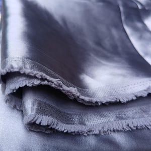 Puede incluir: Primer plano de una tela doblada en tonos azules y grises. La tela tiene una textura sedosa y brillante con un borde deshilachado. Los pliegues crean sombras y reflejos.