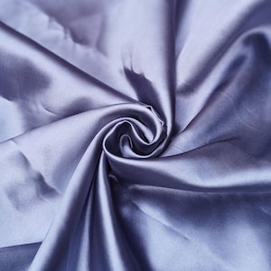 Puede incluir: Primer plano de un remolino de tela de color lavanda. La tela tiene una textura suave y brillante, con pliegues y sombras que crean profundidad. El material parece ser seda o un textil lujoso similar, adecuado para ropa o decoraci&oacute;n del hogar.