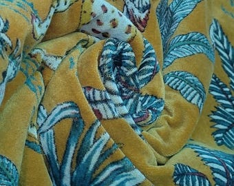Puede incluir: Tela de terciopelo amarillo mostaza con un estampado botánico de hojas y ilustraciones de pájaros turquesas y blancos. La tela es suave y tiene una textura lujosa, con pliegues y sombras que añaden profundidad al diseño.
