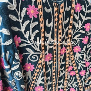 Op de afbeelding: Close-up van een teal jasje met ingewikkelde bloemenborduurwerk. Roze bloemen en grijze bladeren sieren de jas, aangevuld met oranje en beige verticale accenten. Het jasje heeft een hoge kraag en gedetailleerde stiksels.
