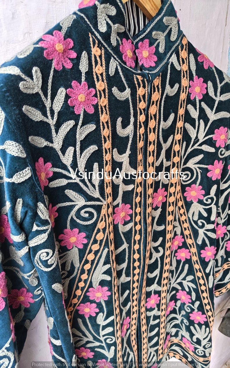 Op de afbeelding: Close-up van een teal jasje met ingewikkelde bloemenborduurwerk. Roze bloemen en grijze bladeren sieren de jas, aangevuld met oranje en beige verticale accenten. Het jasje heeft een hoge kraag en gedetailleerde stiksels.