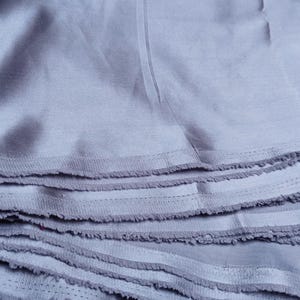 Puede incluir: Primer plano de una pila de tela azul claro con bordes deshilachados. La tela parece ser un material suave y tejido, posiblemente algod&oacute;n o lino. Las capas est&aacute;n cuidadosamente apiladas, mostrando la textura y las variaciones de color.