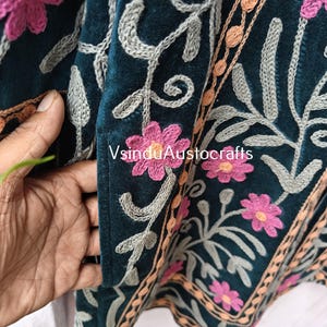 Op de afbeelding: Close-up van een teal stof met ingewikkelde bloemenborduurwerk. Roze bloemen en grijze ranken zijn op de stof gestikt, met een rand van oranje en bruin borduurwerk. De tekst "VsinduAustocrafts" is zichtbaar.