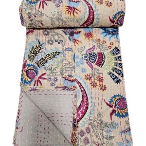 May include: Beige and cream-colored quilt with a floral and paisley design. The quilt features a rolled-up section at the top, showcasing the intricate patterns in shades of pink, blue, and yellow. The edges are finished with a contrasting stitch.
