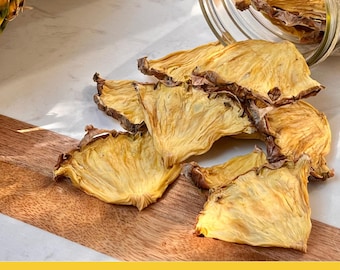 May include: Dried pineapple slices in a glass jar on a wooden cutting board. The pineapple slices are a light yellow color and have a slightly wrinkled texture. The jar is clear glass and has a metal lid. The cutting board is made of wood and has a light brown color. The text "CRAFTED BY THE PERFECT GARNISH CO." is printed on a yellow banner below the cutting board.