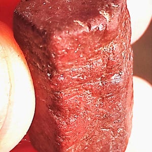 Op de afbeelding: Close-up van een donkerrode, rechthoekige edelsteen. De steen heeft een ruw, getextureerd oppervlak met zichtbare strepen en een licht afgeronde bovenkant. De kleur is een diep, rijk rood.