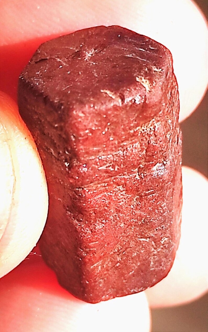 Op de afbeelding: Close-up van een donkerrode, rechthoekige edelsteen. De steen heeft een ruw, getextureerd oppervlak met zichtbare strepen en een licht afgeronde bovenkant. De kleur is een diep, rijk rood.