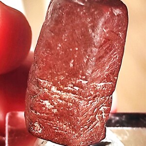 May include: A close-up of a reddish-brown, rectangular stone with a textured surface. The stone has a slightly rounded top and visible white veining patterns. The stone is resting on a white surface.