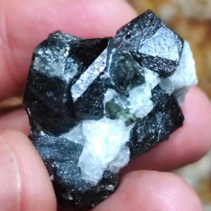 May include: A cluster of black and green crystals with a shiny, metallic finish. The crystals are arranged in a random pattern and have a rough, natural texture.